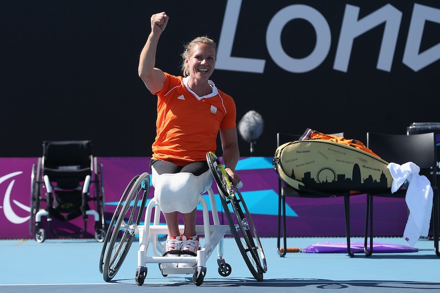 A wheelchair tennis player in an orange shirt pumps her fist in celebration after winning a Paralympic title. 