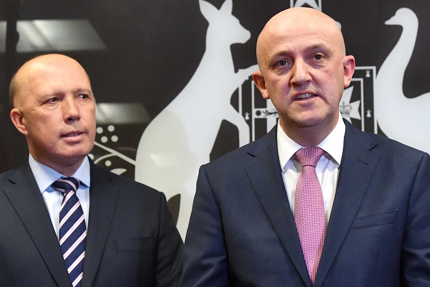 Two men stand at podium during a press conference with the code of arms and the Australian flag in the background