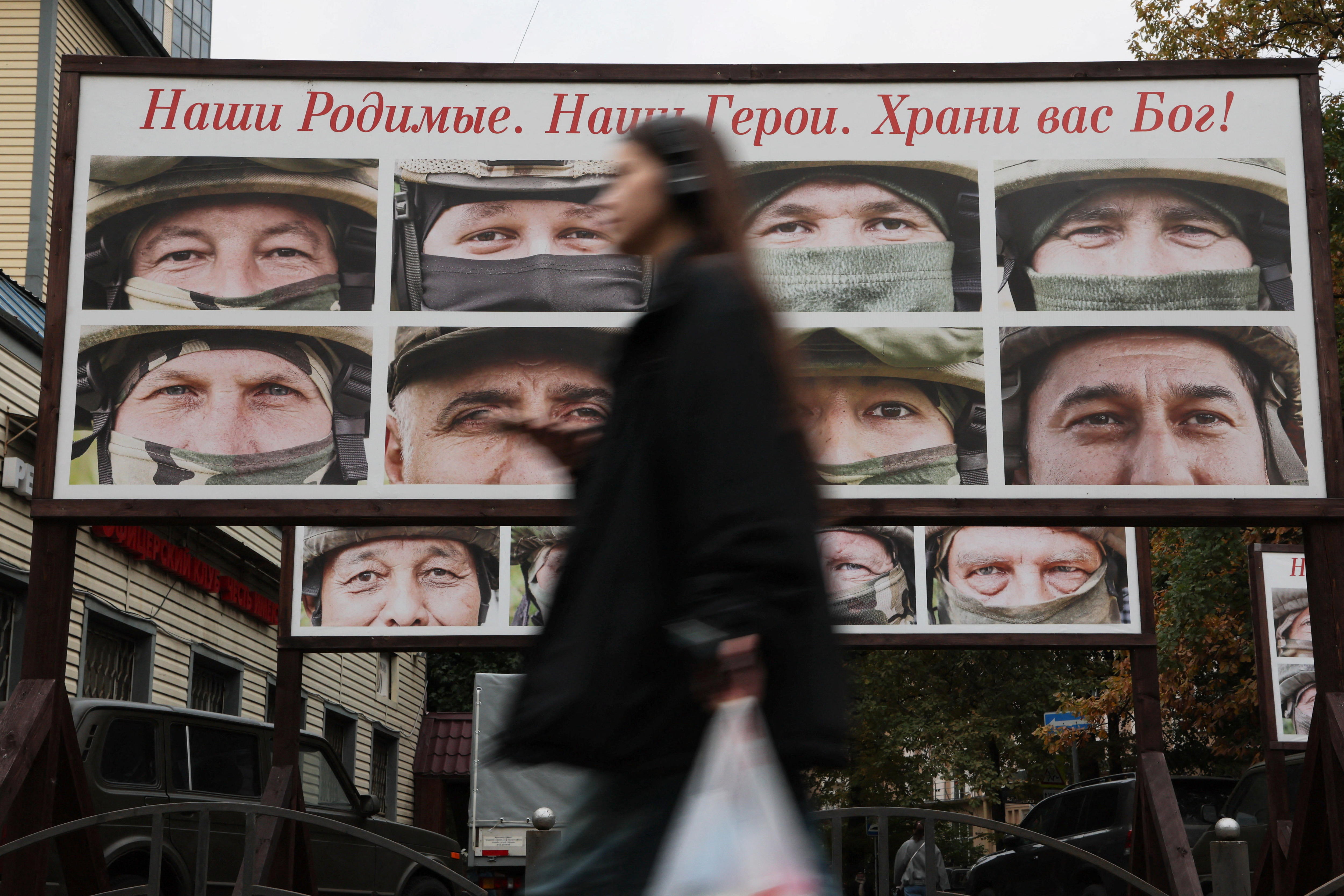 A woman carries grocery bags past a sign featuring close-ups of the eyes of several soldiers.