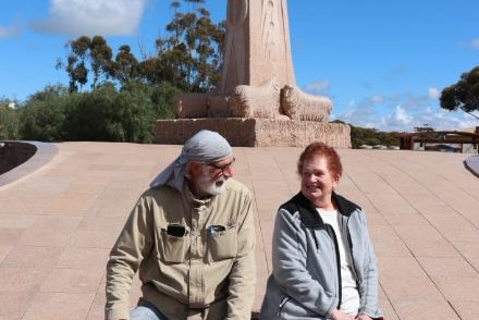 Man and woman sitting on a rock looking at each other in front of large statue featuring sheep at base.