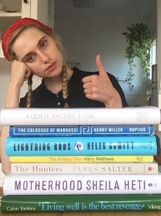 Woman giving the thumbs to the camera, with a stack of books in front of her.