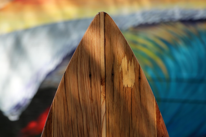 A close-up photo of the tip of a wooden surf board with a surfing mural behind it.