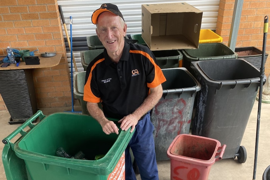 A man stands with a bin and a container with cans and bottles in it. He is smiling and wearing a black and orange shirt.