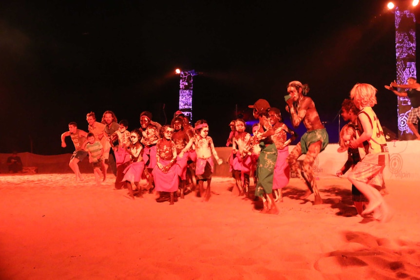 Group of Indigenous and non-Indigenous children and adults dancing together in red dirt at night.