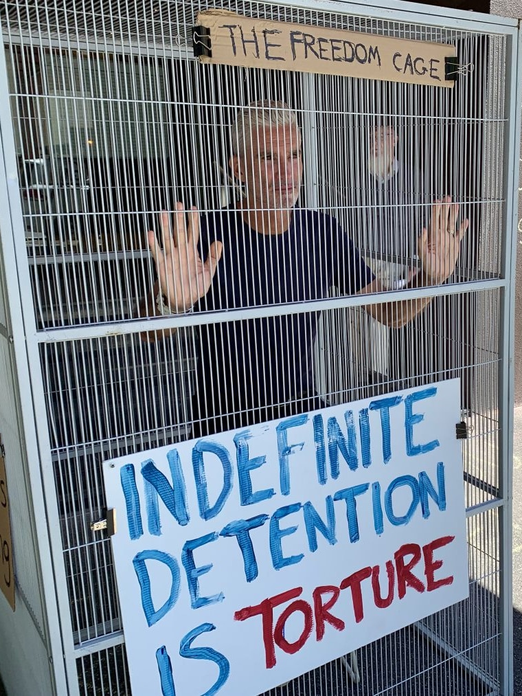 A man in a cage with a sign stating 'indefinite detention is torture'