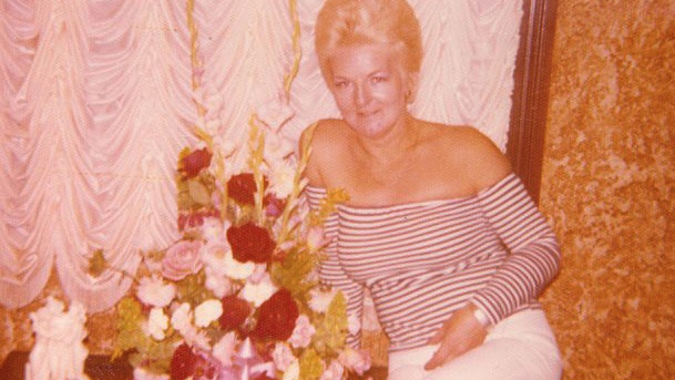 Shirley Finn sits on a small table next to a bouquet of flowers wearing a black and white striped top and white pants.