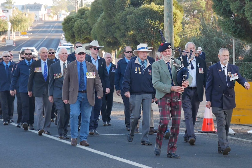 About 20 men led by man playing the bagpipes march along a street in Kojonup.