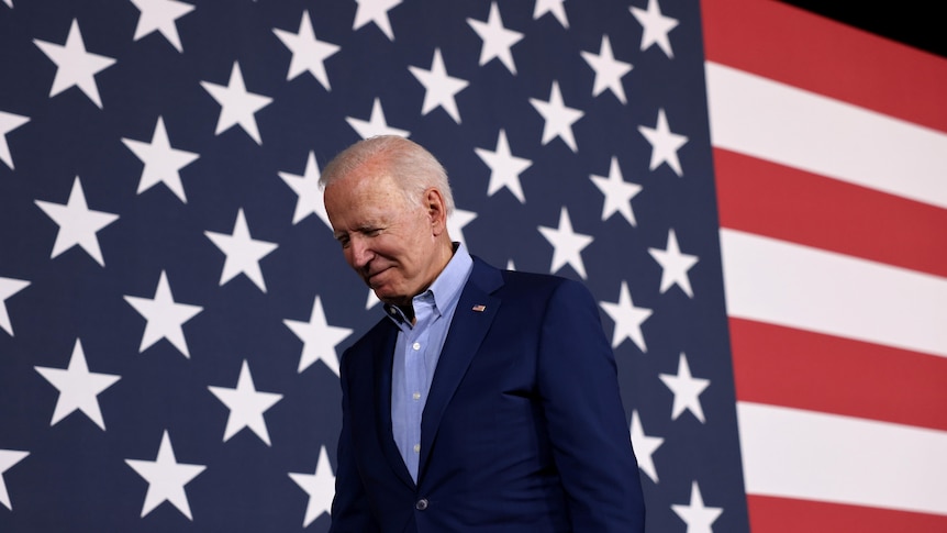 Joe Biden looking down with an American flag behind him 