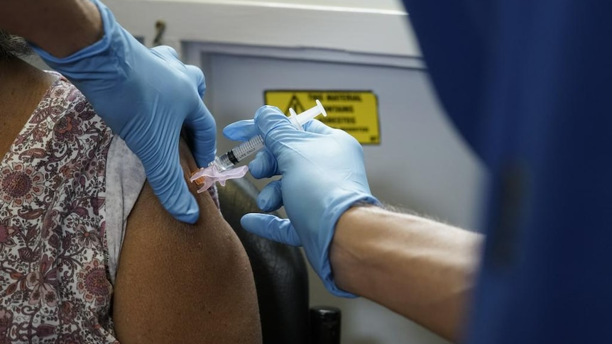 A person's hands in blue gloves, putting a needle into the shoulder of a woman sitting down.