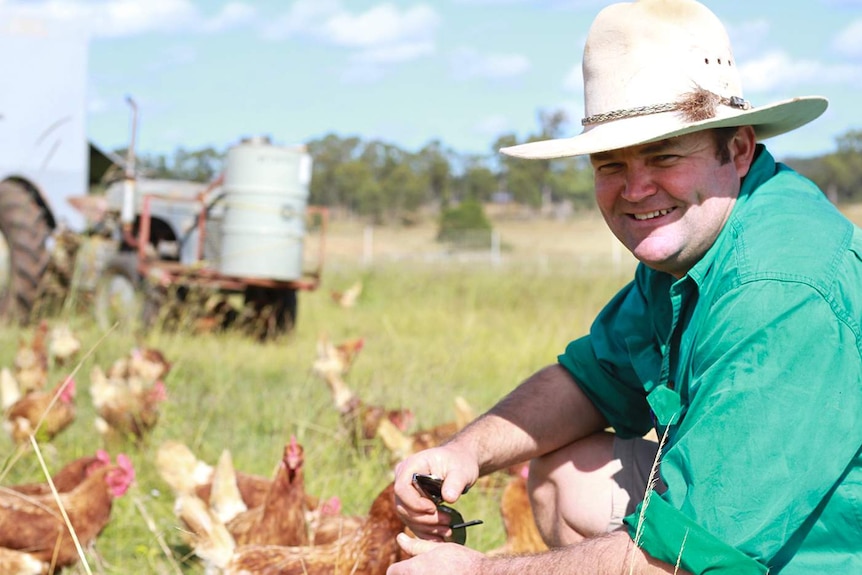 Egg farmer wearing Akubra hat smiles as chooks mill around in a paddock behind him