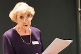 A woman wearing glasses addressing a room with microphone and podium beside her