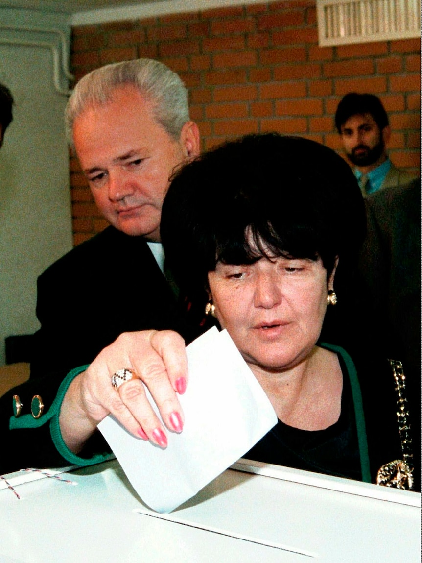 A woman inserts a paper voting ballot into the top slit of a large white cardboard box while a man watches on