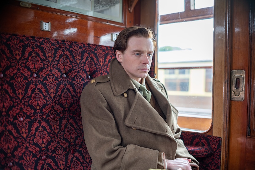 White man with short dark blonde hair wears taupe 1940s trenchcoat in a train carriage with maroon seats.