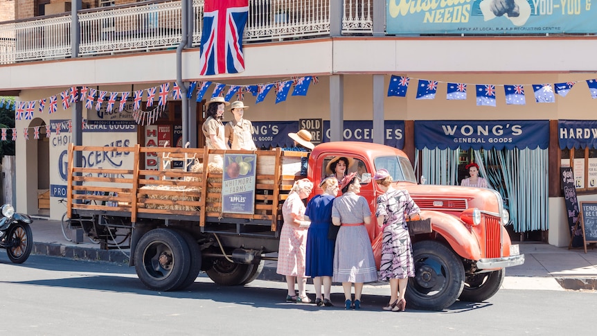 Ladies in 1940s outfits gather round a red farm truck that is parked outside of a small town pub