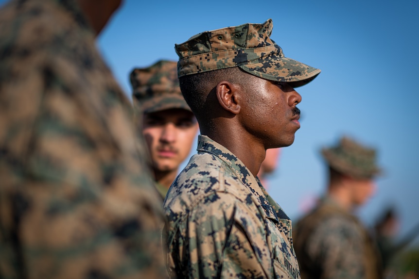 A young male marine with dark skin and a black moustache looks into the distance