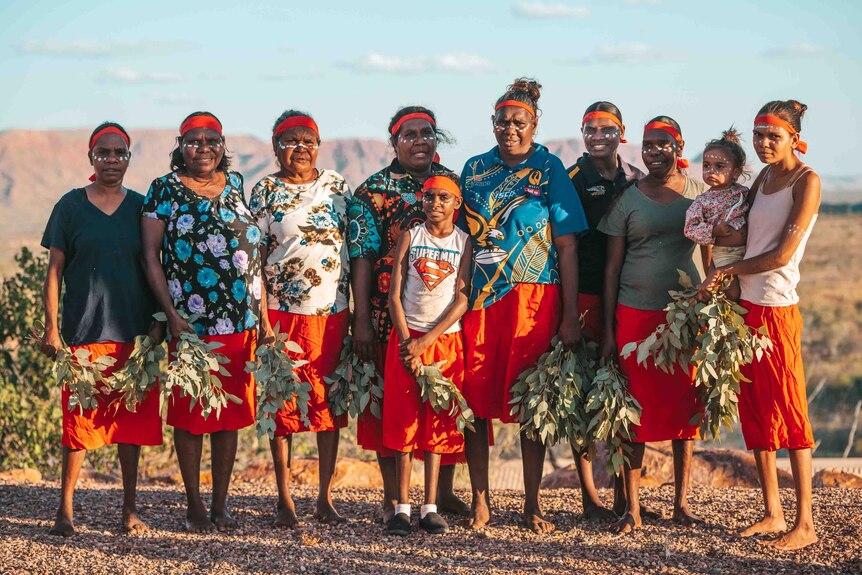 A group of women stand with red headbands and skirts. They are holding bunches of leaves.