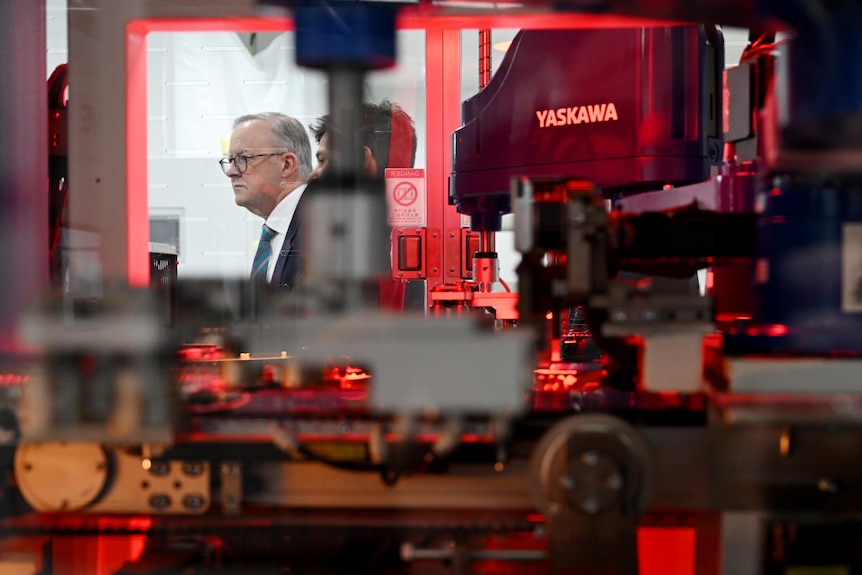 Albanese is seen through the window of a manufacturing facility, production line equipment in the foreground.