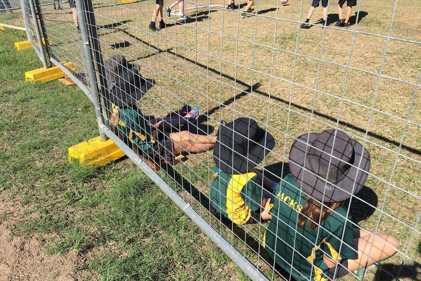 Four children in school uniforms lean against temporary fencing watching other kids play soccer outside