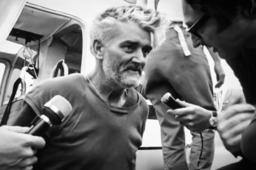 A gaunt looking man disembarks a helicopter to face reporters with microphones.