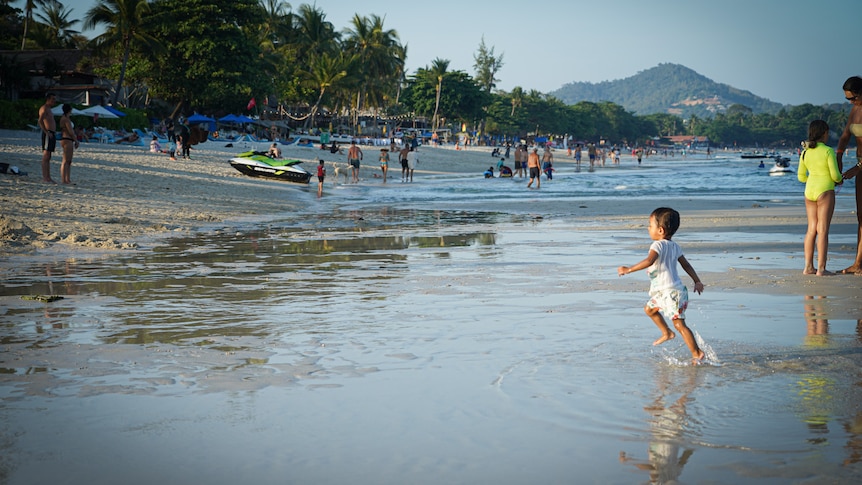A child plays in shallow water on a beach surrounded by other tourists.