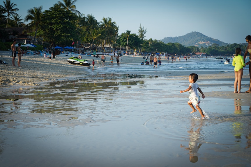 A child plays in shallow water on a beach surrounded by other tourists.