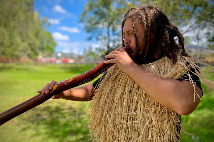 Indigenous man with braided hair wearing paper raffia decoration around his neck and playing the didgeridoo