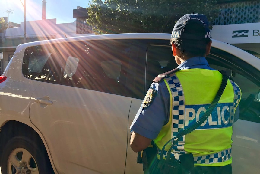 A member of the police force with their back to the camera approaches the driver of a white car to conduct a breath test.