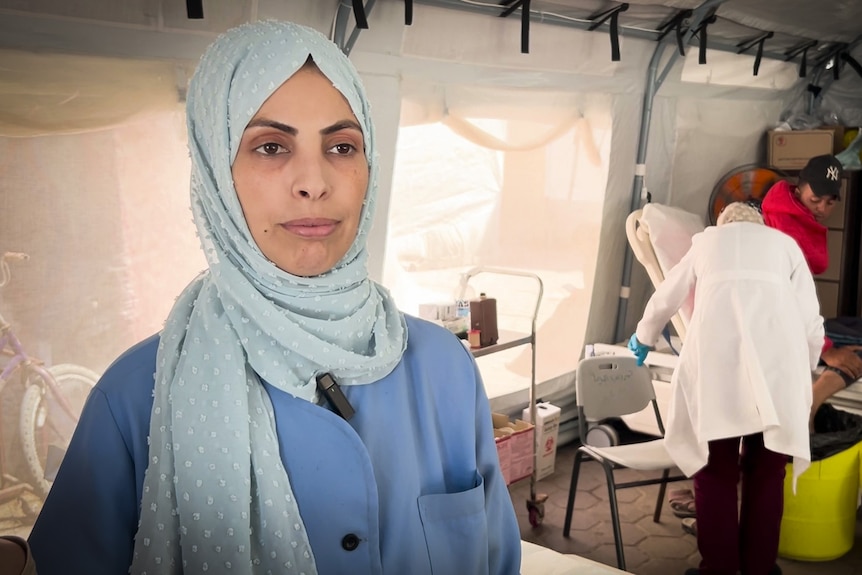 A woman wearing a hijab and blue clothes stands while another doctor tends to a patient.