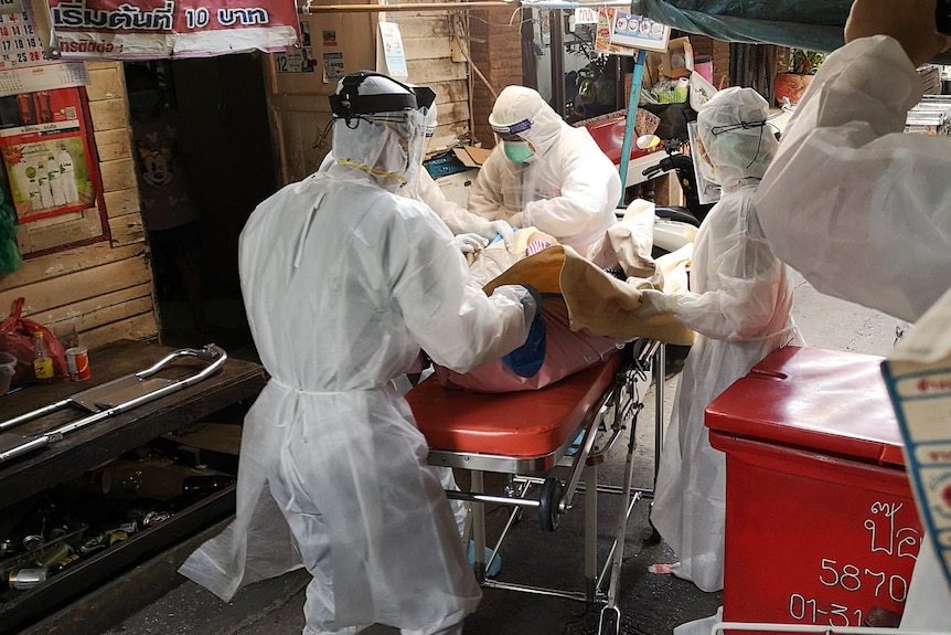 A group of people in head to toe personal protective equipment helf lift a person on a stretcher.