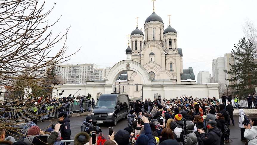 A crowd surround a hearse in front of a church. 