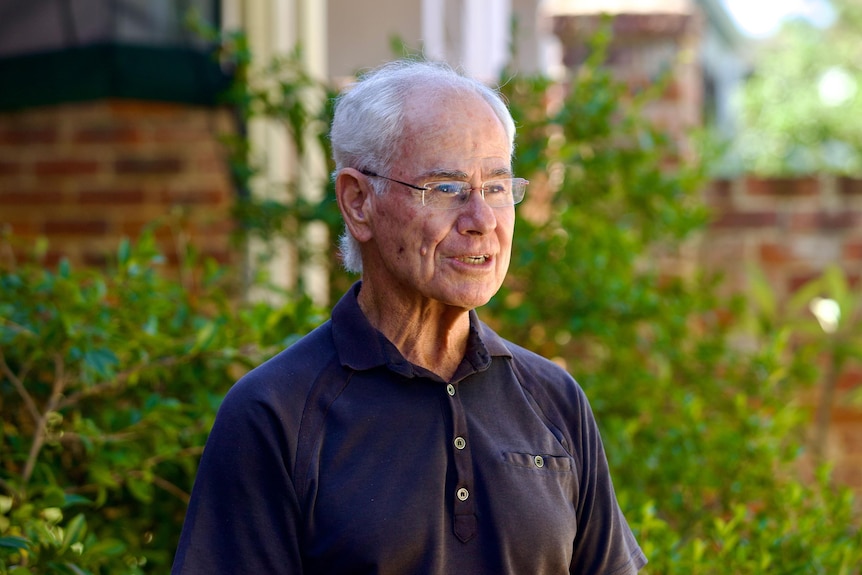 A caucasian man with white hair and glasses looking right while talking. He is wearing a dark grey polo shirt.