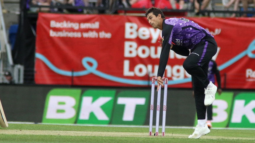 a cricketer in purple uniform is bowling a ball on a cricket field with wickets behind him