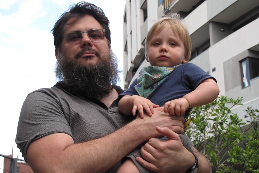 Allister Hill stands in front of a tall building holding his young son in his arms.