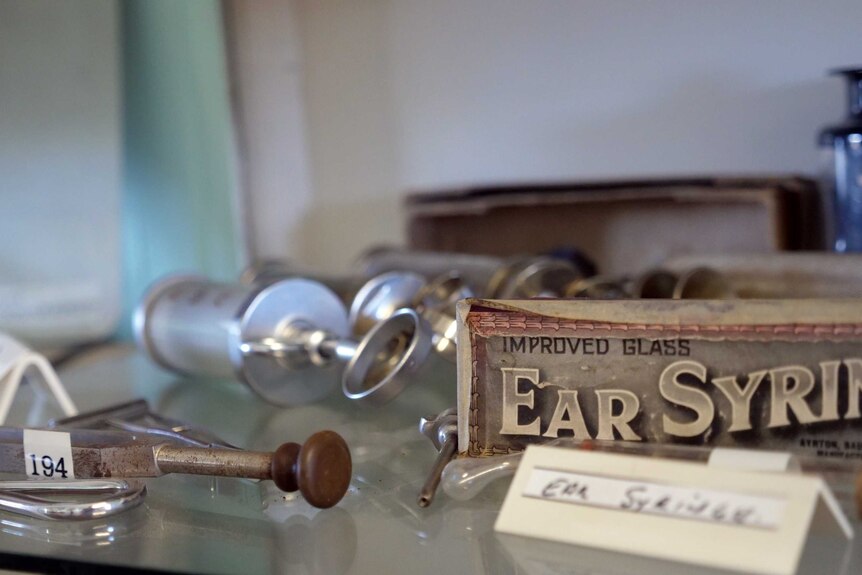 A glass shelf with a collection of old medical instruments. A box labelled ear syringe is visible.