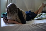 An unidentified teenage girl laying on her stomach on a bed, looking at a mobile phone, with long hair obscuring her face.