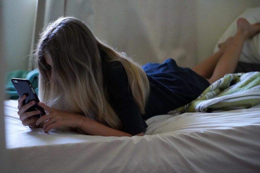 An unidentified teenage girl laying on her stomach on a bed, looking at a mobile phone, with long hair obscuring her face.