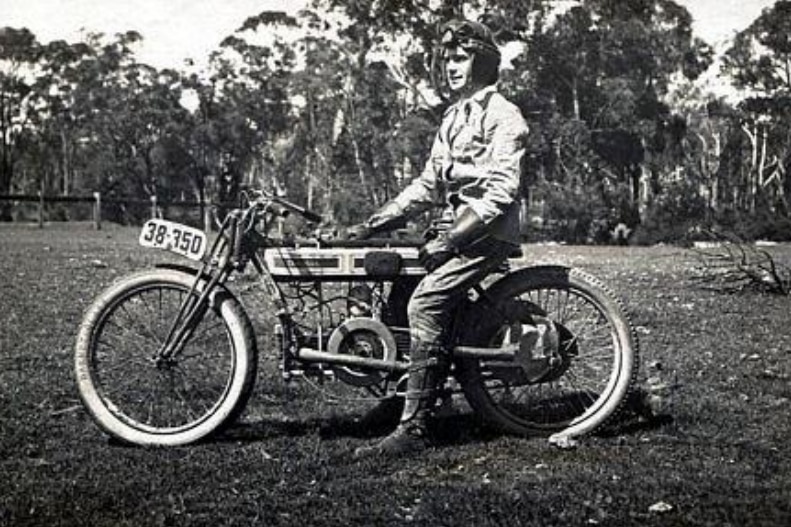 A man sitting on an old motorbike wearing a leather helmet. Image is black and white