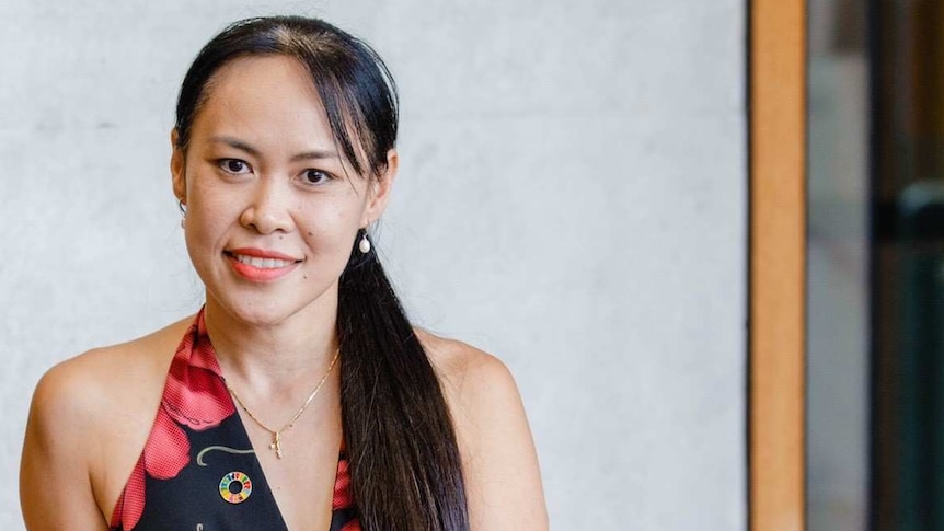 Smiling woman with long black hair in a ponytail, wearing black sleeveless dress with red flowers, sitting on a bench