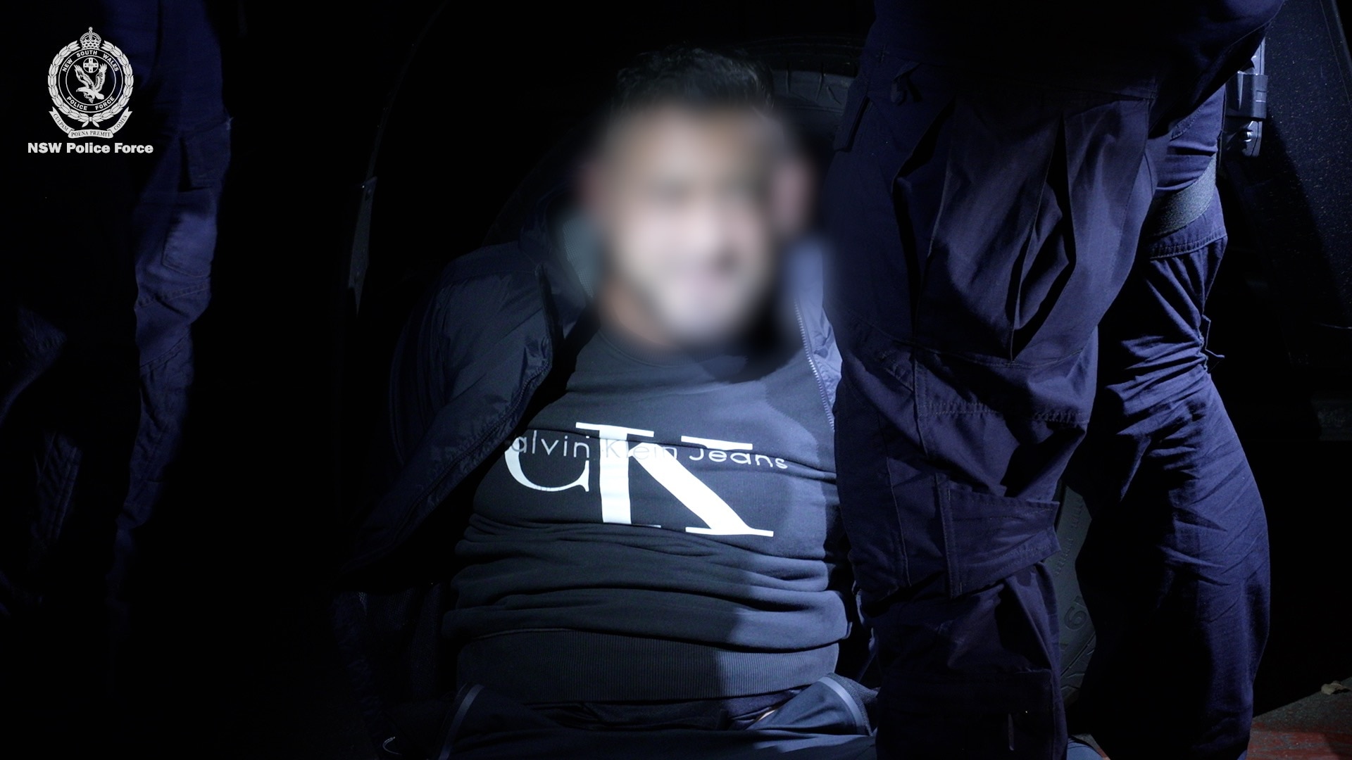 Man in black tracksuit, surrounded by police. Faces blurred