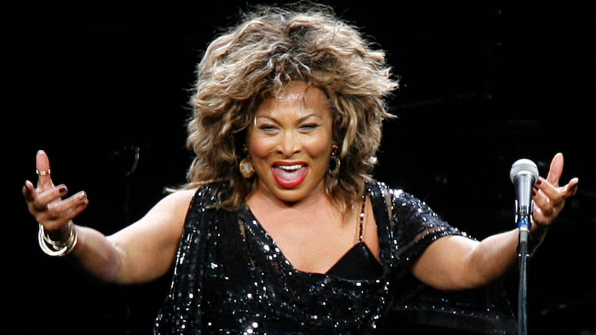 Tina Turner stands behind a microphone and gestures widely, wearing a sparkly black dress.