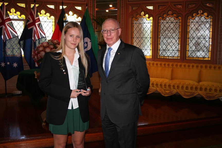 A female teenager in school uniform with a blazer jacket stands next to governor general to receive an award