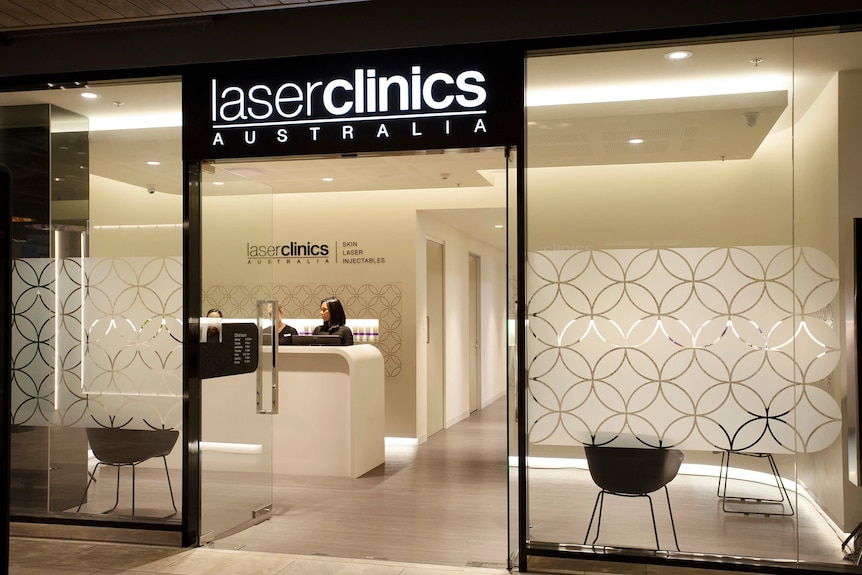 In a soft-lit reception area with sign reading LASER CLINICS AUSTRALIA, two receptionists stand at a desk
