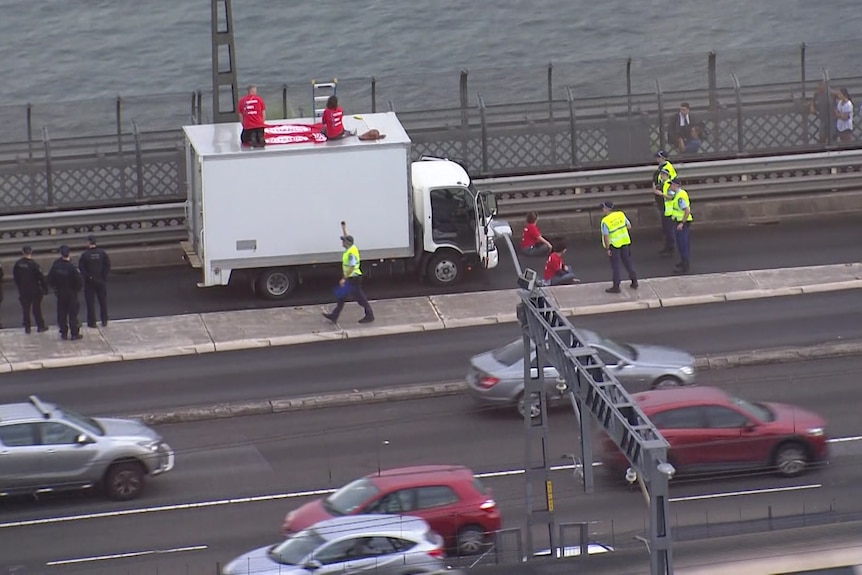 Two people kneel on top of a truck while two sit in front of it as police surround the vehicle