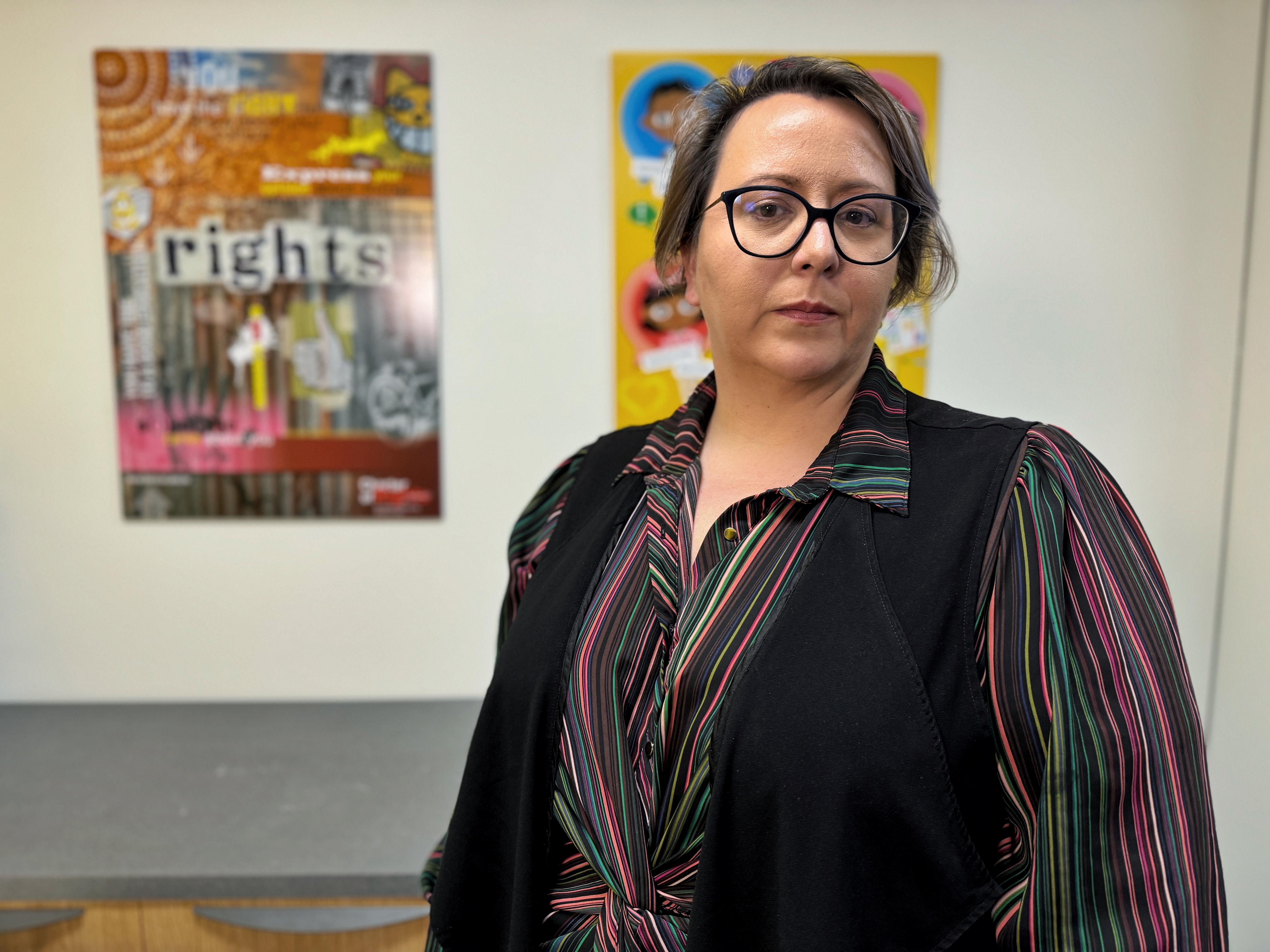 A woman with a serious expression stands in front of posters on a wall