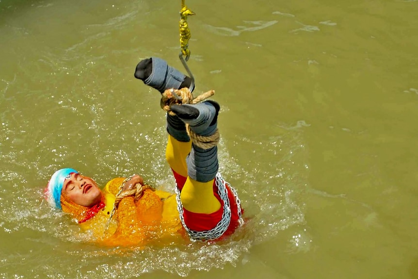 A man wearing chains and ropes is lowered into a river.