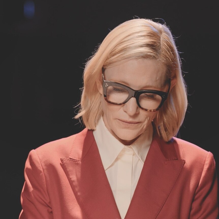 Cate Blanchett wearing a red suit and black glasses looking down at a letter