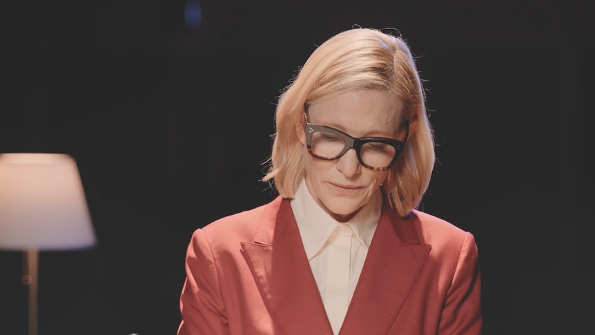Cate Blanchett wearing a red suit and black glasses looking down at a letter