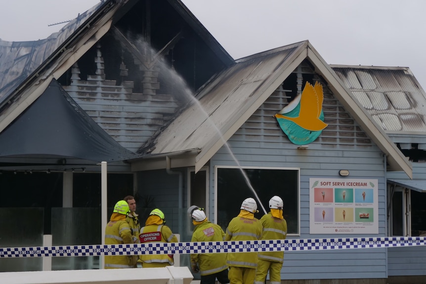 Five firefighters in yellow suits squirting a hose at the roof of a restaurant with police tape in the foreground.
