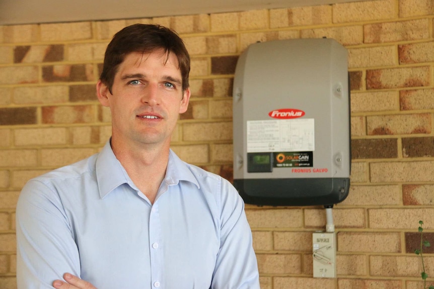 Headshot of Tim Clifford next to a solar energy inverter mounted on a brick wall.