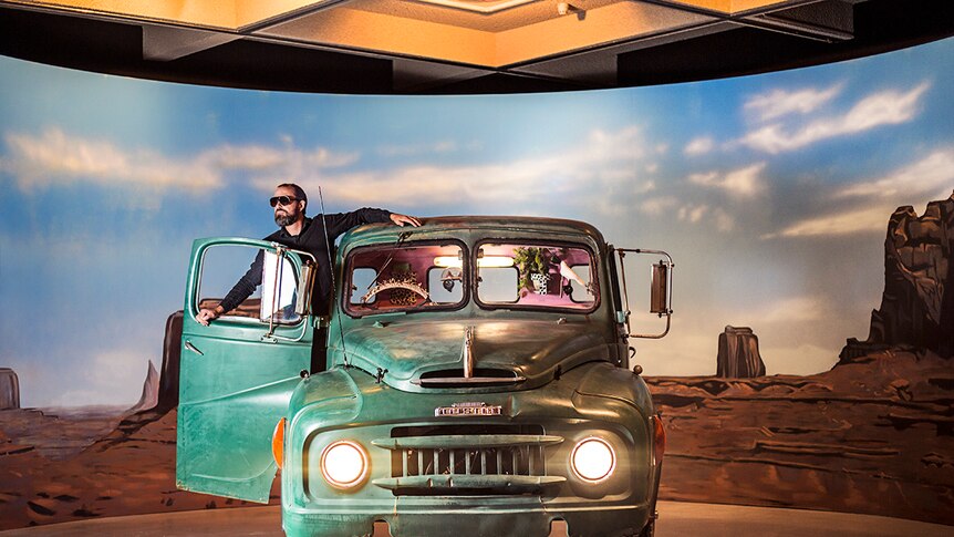 Colour photograph of artist Mark Shorter standing in a green truck in a gallery in front of a painted backdrop.